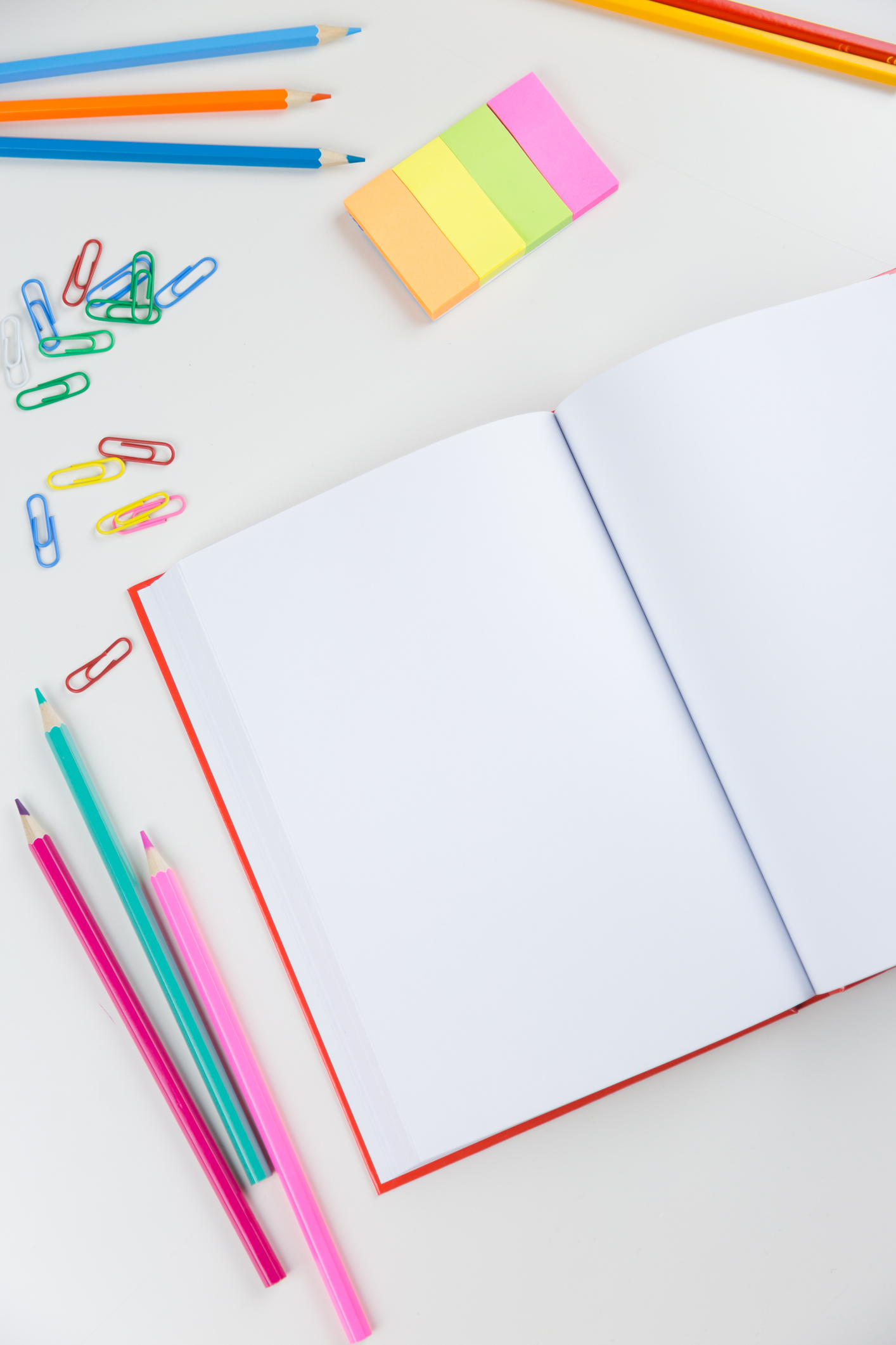 High Angle View Of Book With Colored Pencils And Paper Clips On White Background