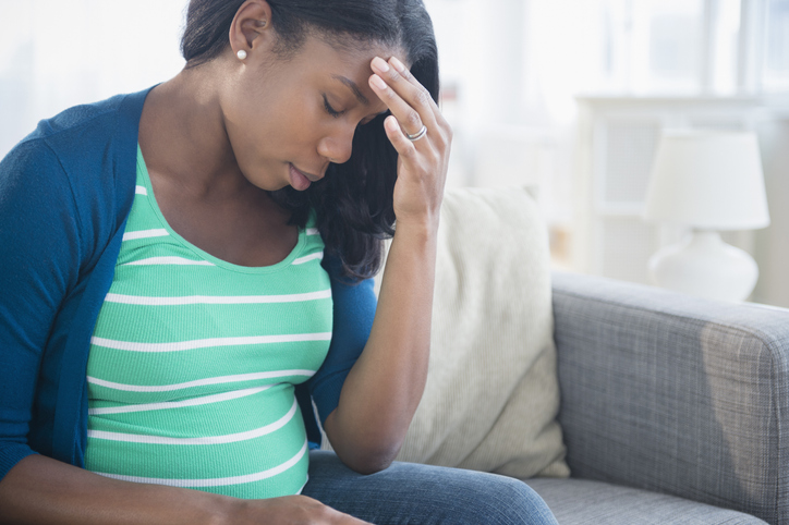 Anxious Black pregnant woman rubbing forehead on sofa