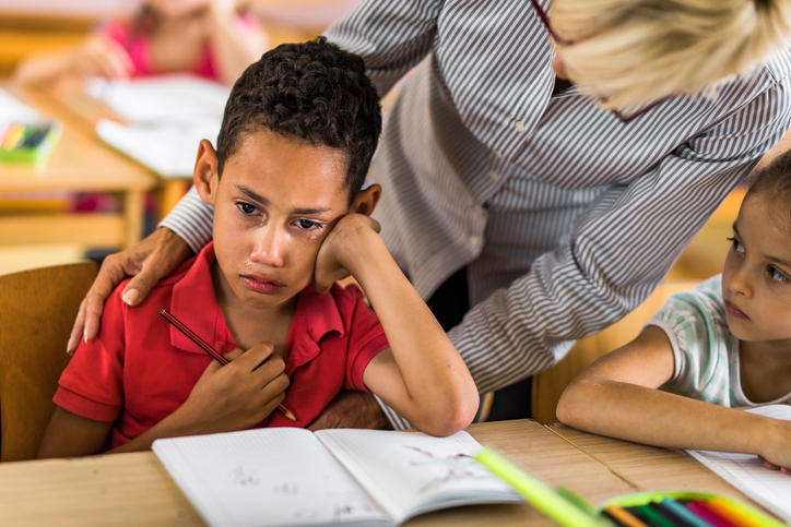 African American elementary student crying on a class while his teacher is consoling him.