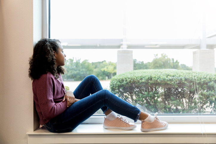 Sad young girl sitting on windowsill looking out window