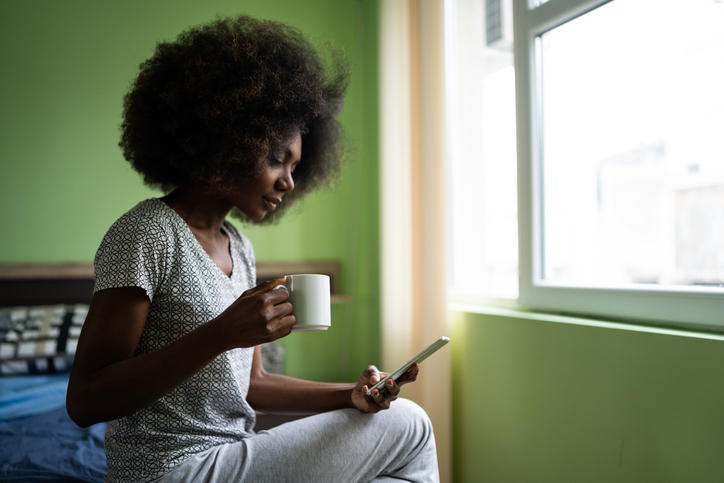 Afro woman sitting on a bed using smart phone and drinking coffee