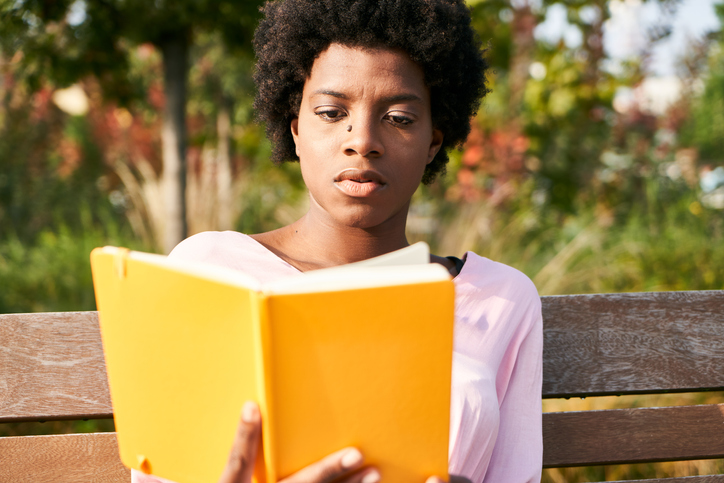 Young Woman With Afro Hair Reading Book While Sitting On Bench