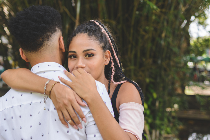 Portrait of young afro couple hugging in a public park. Rear view of the man. Woman looking at camera over the her boyfriend's shoulder.