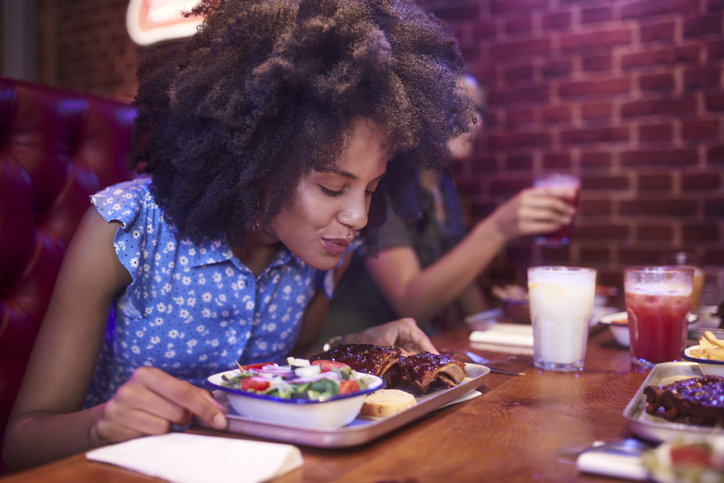 Close-up of african-american woman enjoying and smelling bbq ribs in retro restaurant with friends
