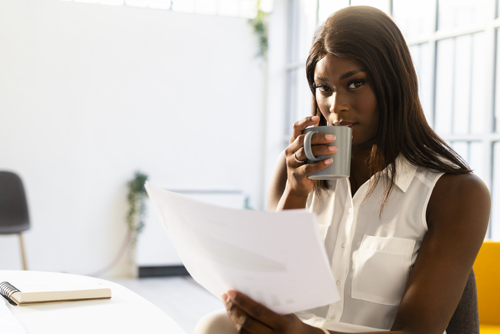 Young woman with coffee cup reading paper while sitting on chair at office
