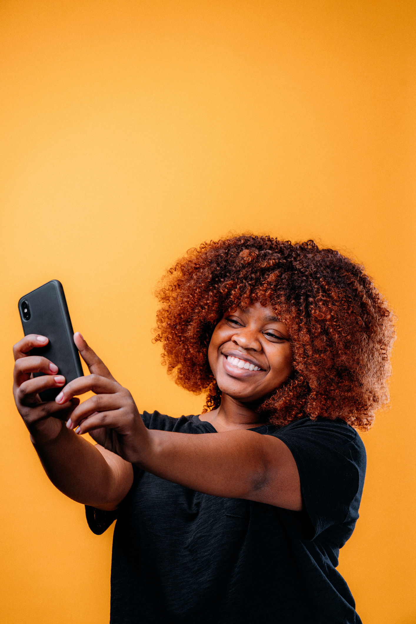 Studio Portrait of a Confident, Beautiful Young African American Woman Using a Mobile Handheld Smartphone to Snap a Selfie Photo for Social Media in Front of a Happy Mustard Yellow-Colored Background
