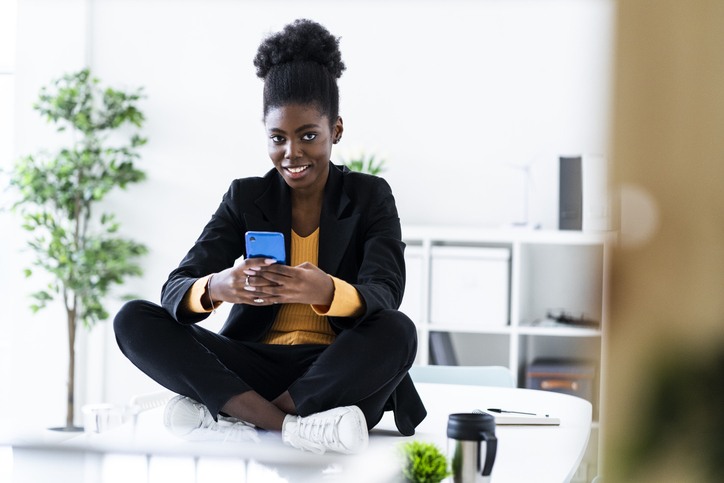 Smiling young Afro female professional text messaging using mobile phone while sitting cross-legged on desk
