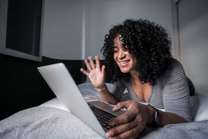 Young woman waving while doing a video call on laptop lying in bed at home
