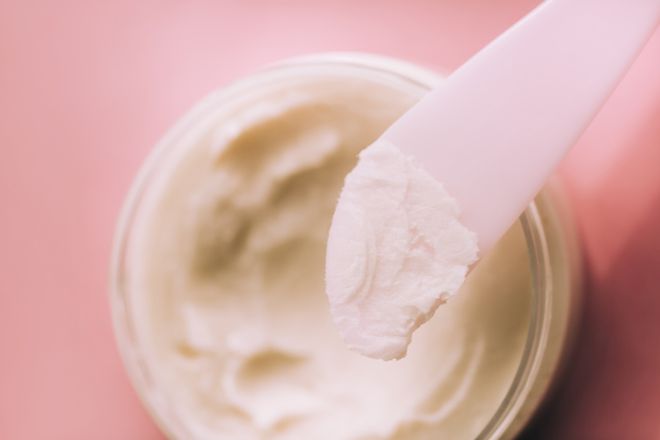The coconut oil on spatula directly above glass jar with coconut oil on pink background. Trendy products of the year. Health and wellness concept