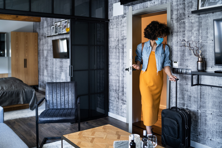 Young afro american woman with protective face mask, holding suitcases entering a rented flat or hotel room