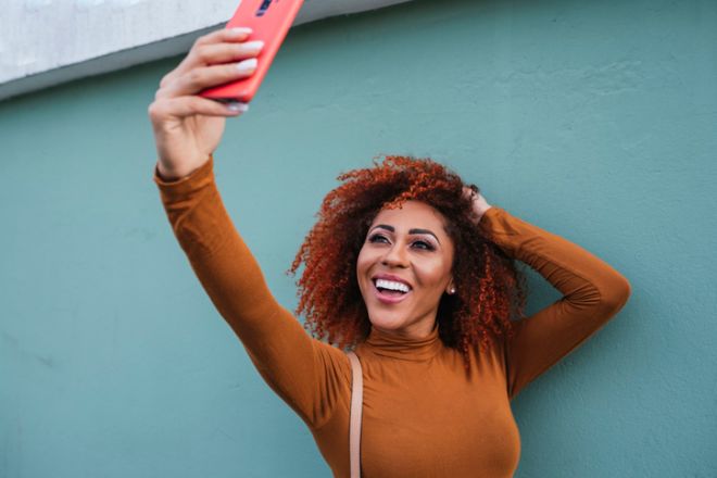 Cheerful afro young woman taking selfie with smart phone while standing by wall