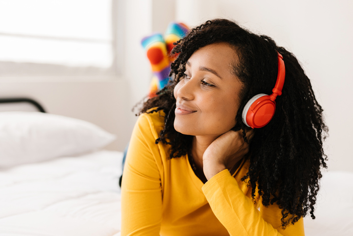 Smiling young brazilian woman with curly hair listening music with headphones while lying on a bed in room