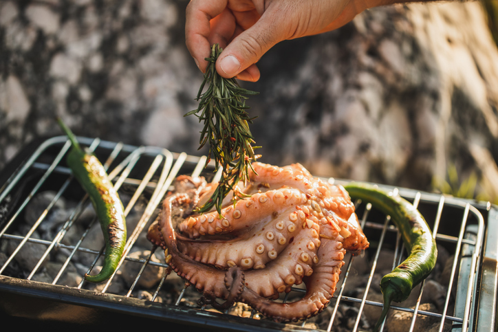 Close up of manâ€™s hand with rosemaryâ€™s bunch cooking octopus on barbecue.