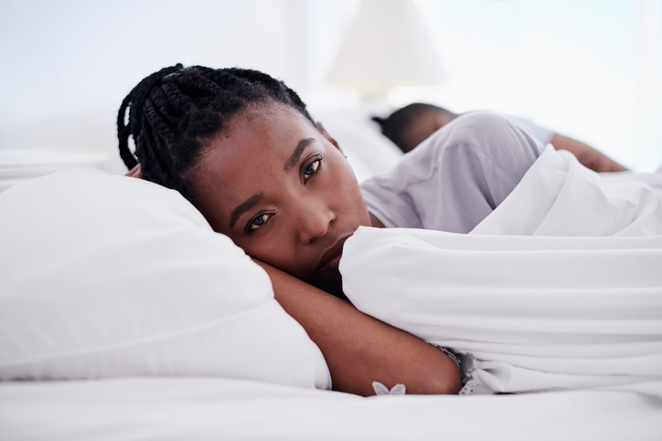 Shot of an attractive young woman lying in bed at home and looking upset while her boyfriend sleeps behind her