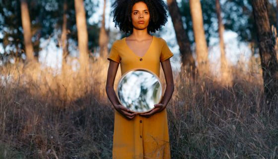 Young woman with afro hair holding mirror while standing in forest at sunset