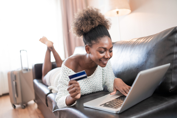 Smiling african woman lying on the couch and using credit card and laptop
