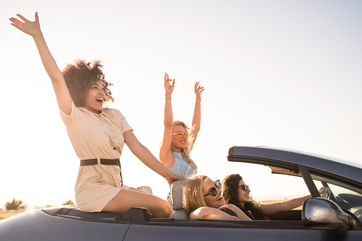 Side view of multiracial group of excited young women enjoying a car ride in a convertible car on a sunset