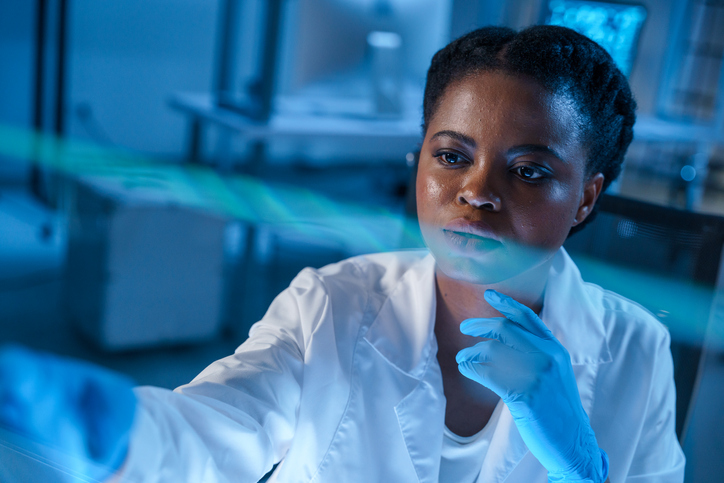 A thoughtfully looking away African - American female doctor in white lab coat in a modern laboratory at her workplace, we see her in close-up in a blue color scheme