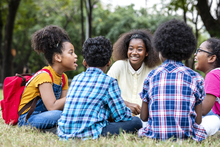 Group of elementary children sitting in a circle on the grass while talking, playing together at a park. Outdoor classroom.
