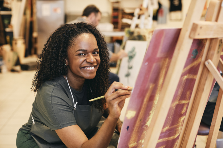 Portrait of smiling mature woman with paintbrush sitting by easel in art class
