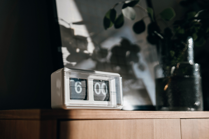 A white digital flip clock shows six o'clock on a brown wooden cabinet next to a fresh eucalyptus plant in the living room, with sunbeam shining through the window on a fresh beautiful morning. A brand new day, fresh start, fresh energy, new opportunities