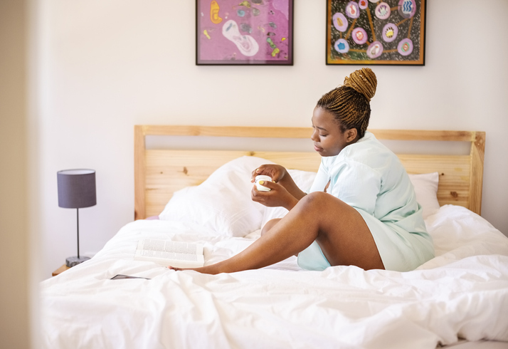 Young african woman with cream for feet sitting on bed in morning