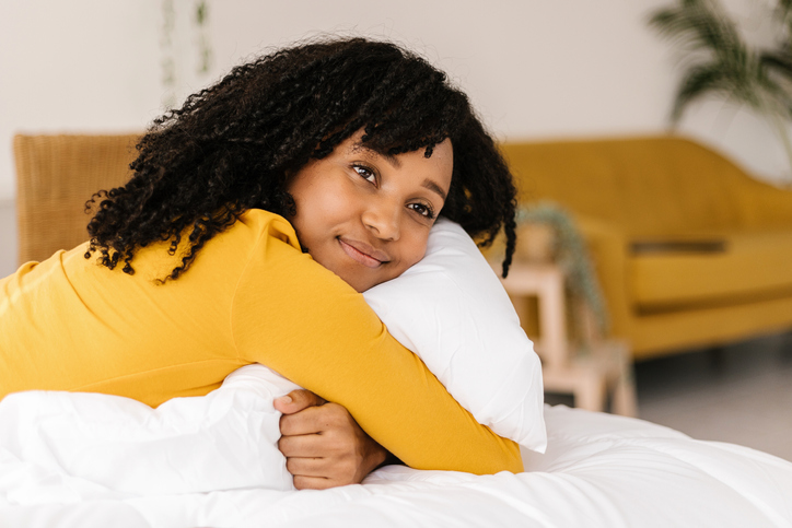 Young dreamy african american woman hugging a pillow while lying on a bed at home.