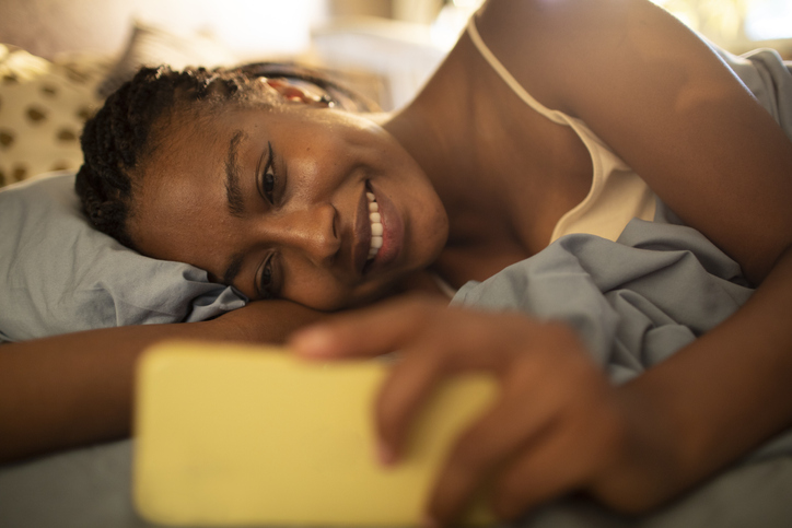Young African woman using her phone while lying down in bed