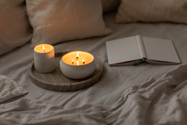Scented candles in ceramic bowls on linen bed with book at home.