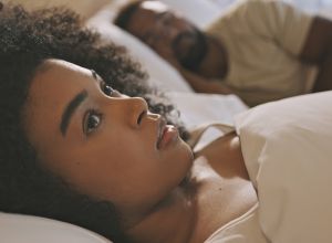 Worried black woman laying in bed with insomnia looking anxious and concerned, having infidelity and relationship issues. Man sleeping while his wife lays awake at night feeling depressed or troubled