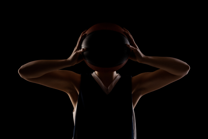 Female basketball player. Beautiful girl holding ball in front of face. Side lit silhouette studio portrait against black background..