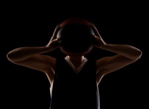 Female basketball player. Beautiful girl holding ball in front of face. Side lit silhouette studio portrait against black background..