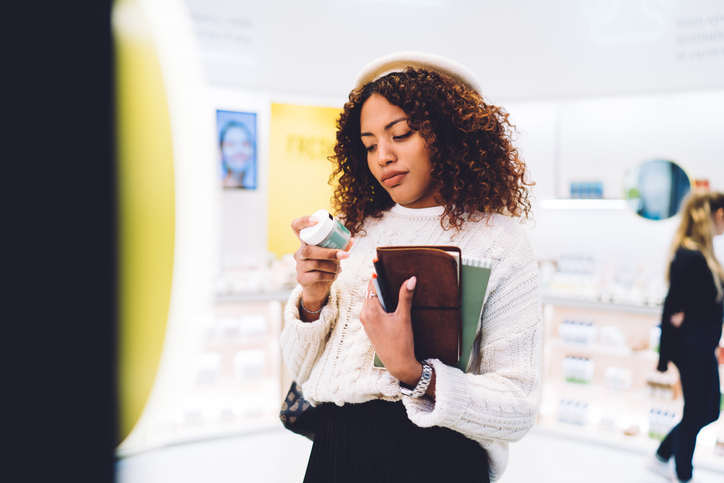 Pensive trendy dressed african american woman checking ingredients and chemicals on cream standing in pharmacy for buying cosmetics, beautiful dark skinned hipster girl selecting product in store