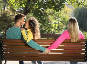 Man holding hands with another woman behind his girlfriend's back on bench in park. Love triangle involved in mate poaching
