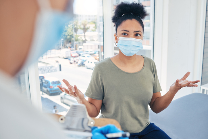 Covid, healthcare and frustrated patient in a hospital with a doctor annoyed at a delay during a medical appointment for consulting and advice. Woman in a mask feeling upset during medical checkup