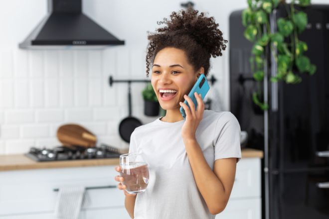 Healthy lifestyle concept. Smiling African American woman holding glass of clean water, talking on mobile phone while standing in kitchen at home