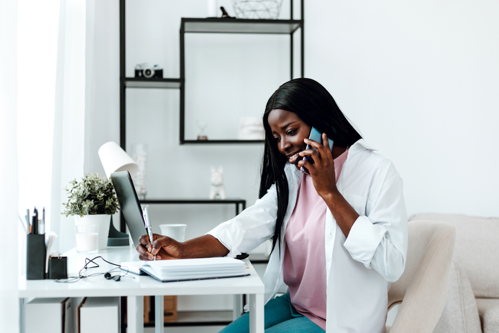 Beautiful young Black woman talking by mobile phone while working at home.
