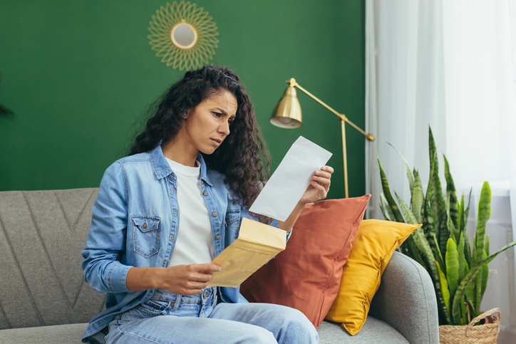 Young beautiful hispanic woman at home disappointed and sad received letter from university about bad exam results, student with curly hair sitting on sofa in living room