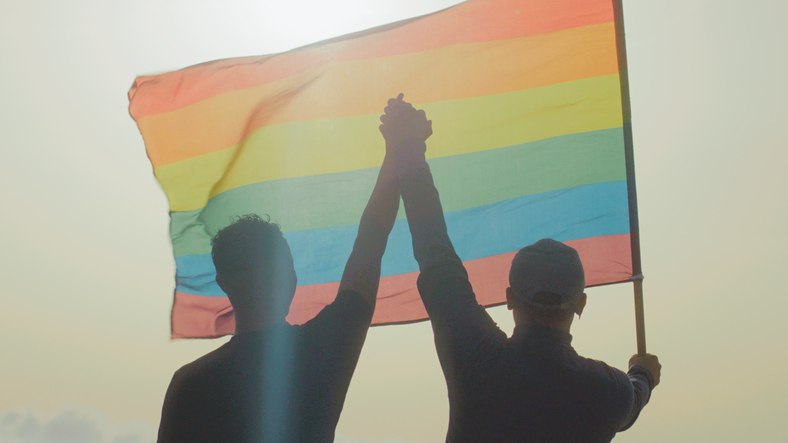 Two gay man celebrating LGBT pride month. Holding hands in air and waving rainbow flag celebrating Respect for Marriage Act