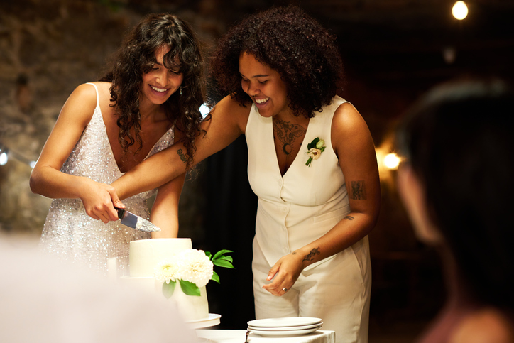 Two young cheerful brides in elegant wedding attire cutting big cake