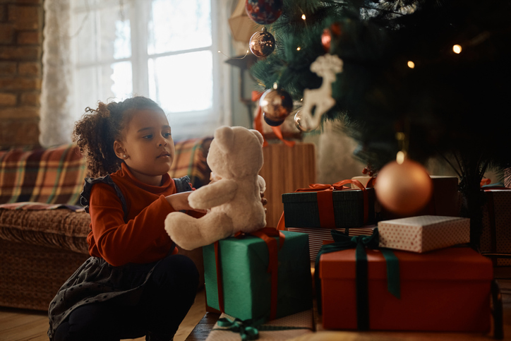 Little black girl playing with plush toy by Christmas tree at home.
