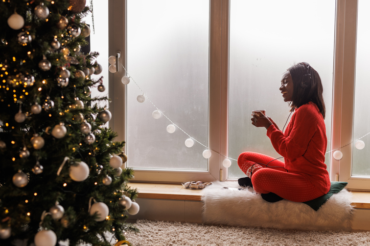 Cheerful young woman relaxing on the window sill, listening to music and enjoying a hot beverage and Christmas activities