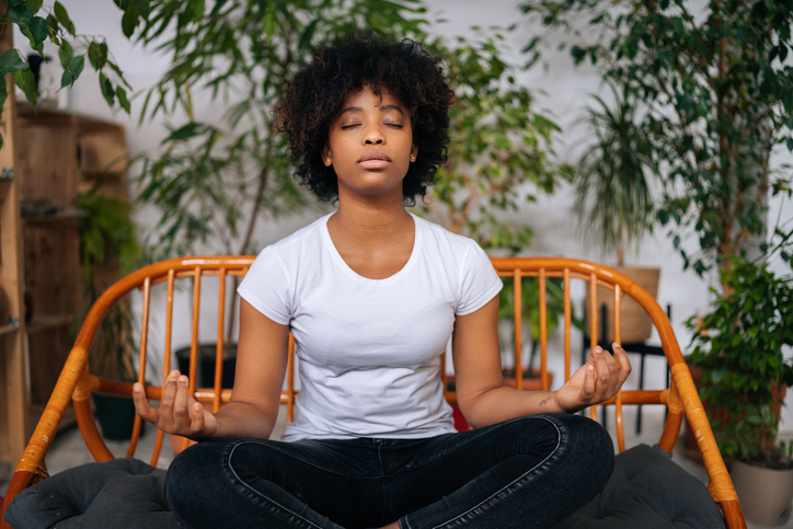 Portrait of peaceful black young woman sitting with closed eyes on sofa in lotus pose, practicing zen energy through yoga meditation at home, relaxing and breathing deep.