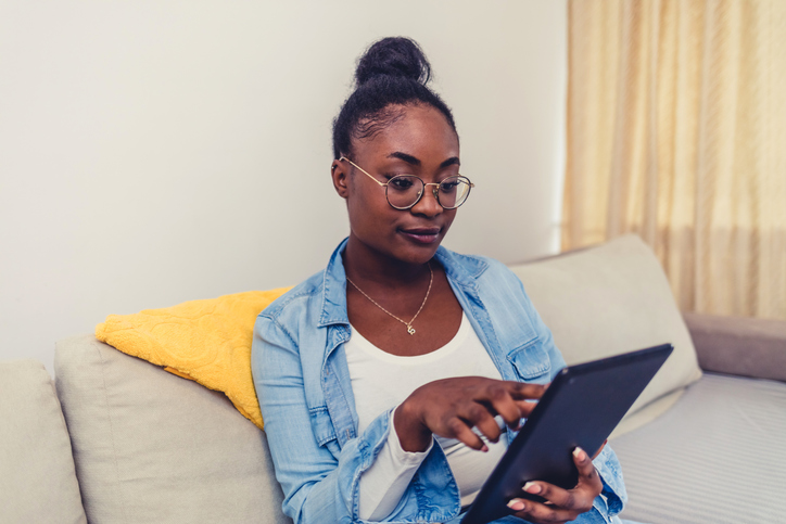 Shot of a young woman using a digital tablet on the sofa at home. Side view of African-American woman sitting on the couch and looking at a digital tablet.