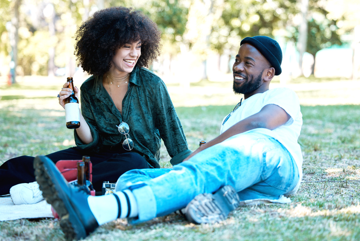 Interracial couple in nature park together, man and woman relax with drink on grass and happiness for people on an inflation friendly picnic date outside. Happy black friends dating, laughing and having fun on weekend