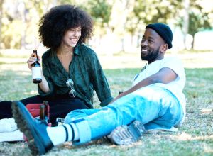 Interracial couple in nature park together, man and woman relax with drink on grass and happiness for people on an inflation friendly picnic date outside. Happy black friends dating, laughing and having fun on weekend