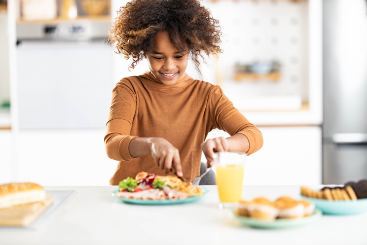 Happy African American girl cutting meal in dining room.