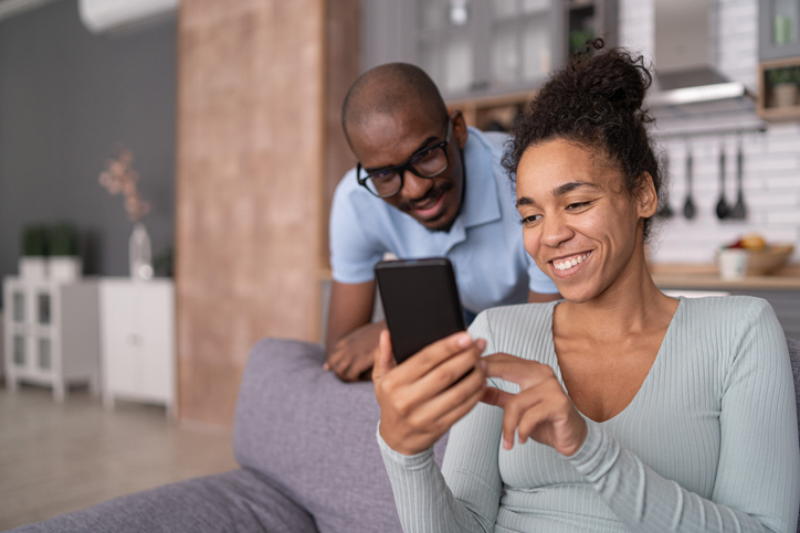 Happy young African American family couple using cellphone, laughing watching funny video online, online video calls, sitting on the sofa at home. Woman showing her mobile screen to her man.