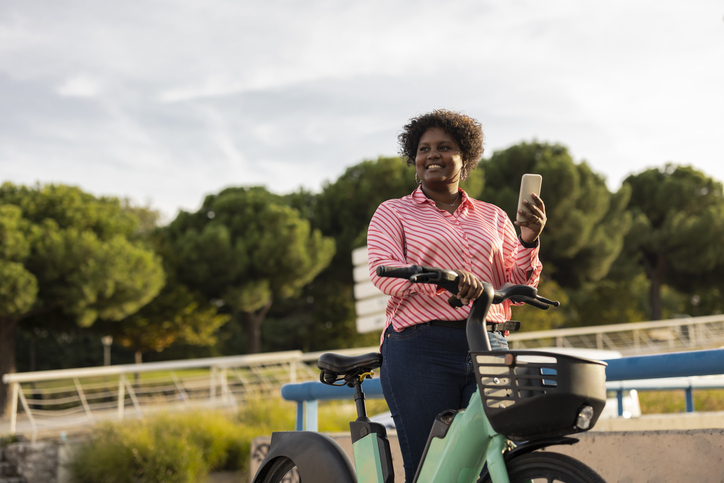 African curvy woman with stylish curly hair on an electric bicycle with smartphone in hand