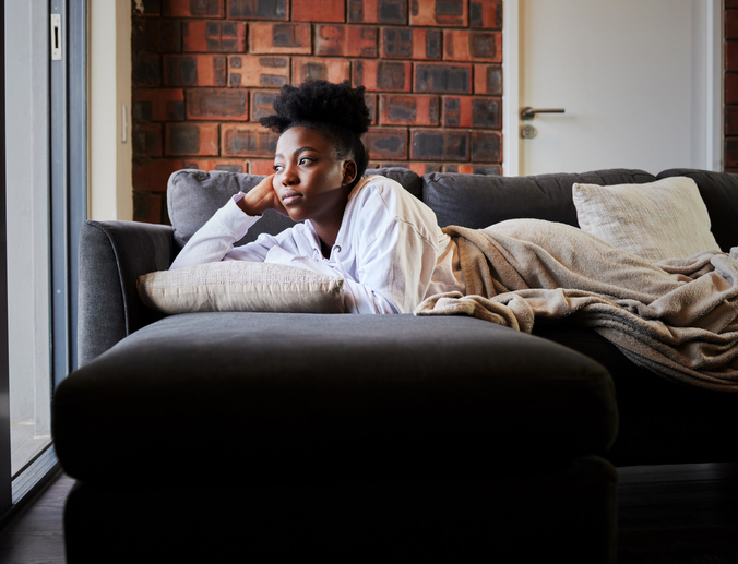 Bored, depression and isolation with black woman lying on her sofa during lockdown, quarantine or pandemic. Mental health, anxiety and stress with young female thinking and looking out her window